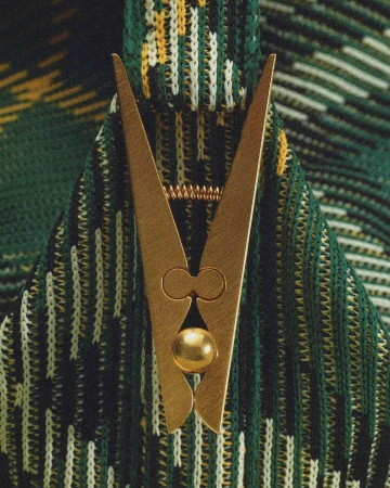 Close-up of a brass-colored clothespin-shaped decorative object against a green, yellow, and white knitted background.