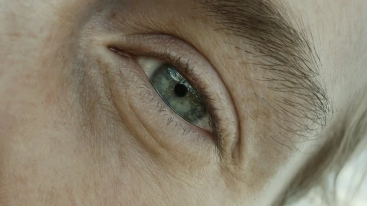 Close-up shot of a person's eye with blue and green hues, capturing detailed textures of the surrounding skin and brow.