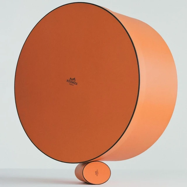 Stacked orange round boxes with the Hermes logo create a minimalist composition against a neutral backdrop in a studio setting.
