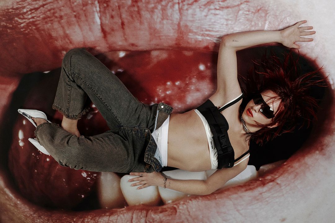 A fashionable young woman with vibrant red hair, wearing stylish jeans, a bra top, and sunglasses, lies in a mouth-themed backdrop.