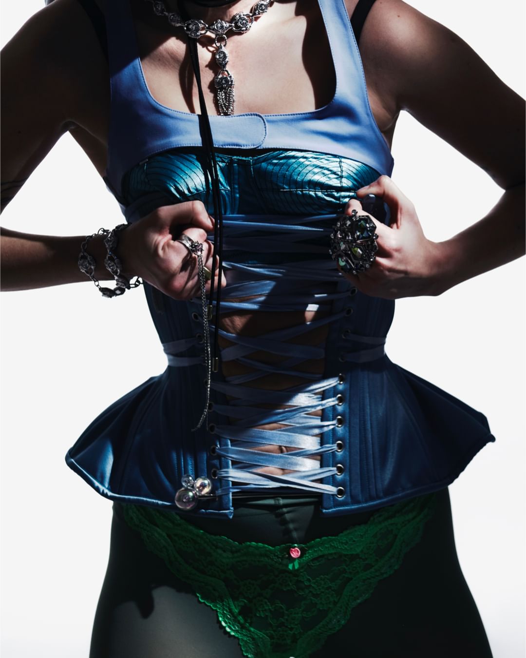 Close-up of a woman wearing a blue corset, elaborate silver jewelry, and green lace underwear against a bright, neutral background.