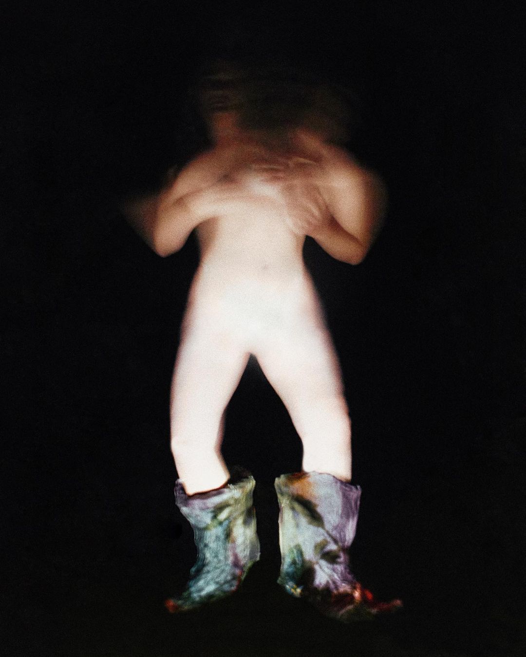 Abstract studio shot of a nude figure wearing a pair of multi-colored boots against a dark background with motion blur.
