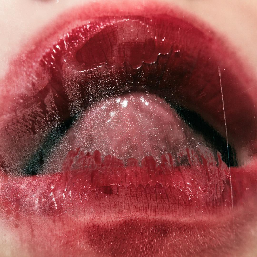 Close-up photograph of a woman's open mouth with a visible tongue, wet teeth, and glossy, dark red lipstick on her lips.