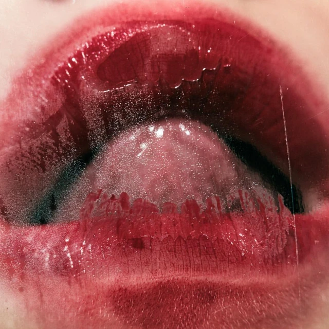 Close-up photograph of a woman's open mouth with a visible tongue, wet teeth, and glossy, dark red lipstick on her lips.
