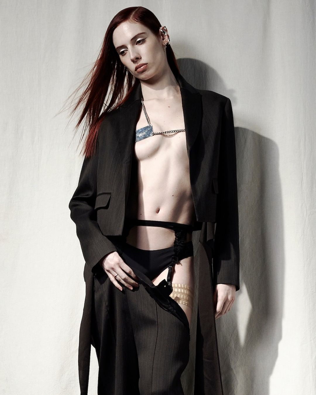 A slender redhead fashion model in a dark pin-stripe coat poses indoors against a cream-colored wall, showing off an edgy look.