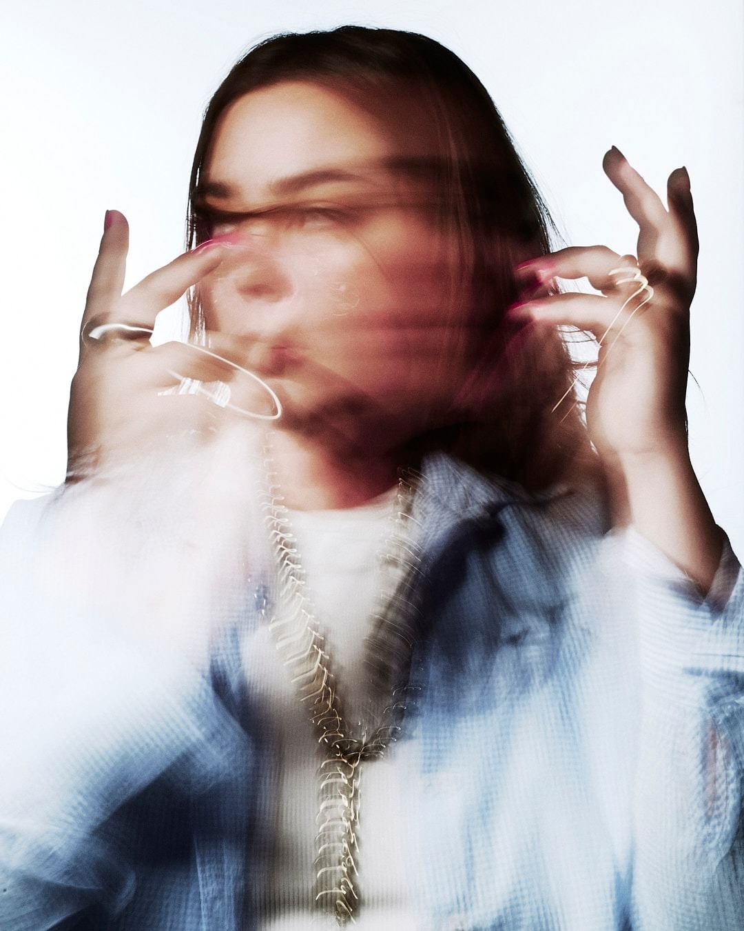 Portrait of a woman with motion blur effect wearing a blue button-down shirt and layered metallic necklaces against a white background.