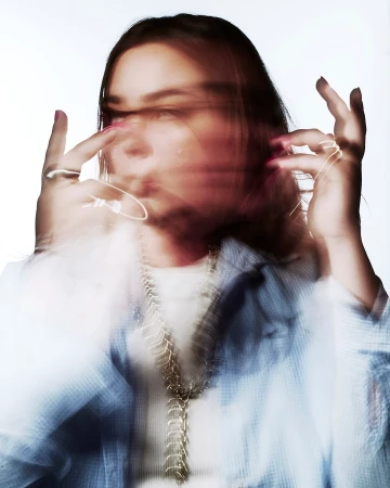 Portrait of a woman with motion blur effect wearing a blue button-down shirt and layered metallic necklaces against a white background.