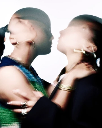 Close-up of two women with blurred motion, face to face with gold jewelry. The blurred motion gives a sense of movement and energy.