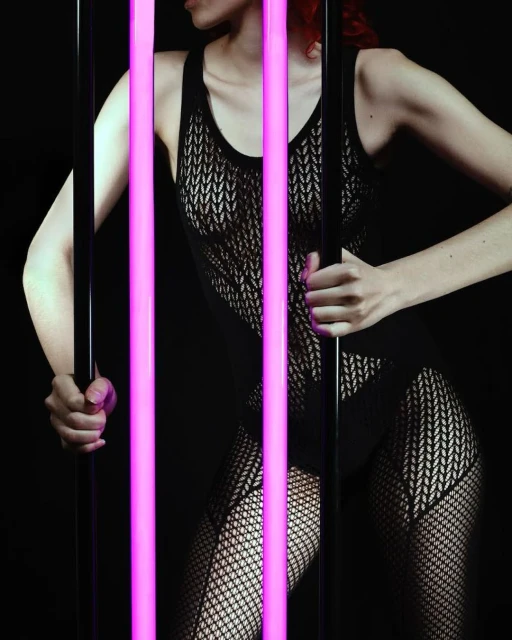 A woman poses in a black, sheer bodysuit and fishnet stockings, standing behind vertical neon tubes in a dark photo studio setting.