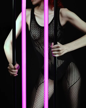 A woman poses in a black, sheer bodysuit and fishnet stockings, standing behind vertical neon tubes in a dark photo studio setting.