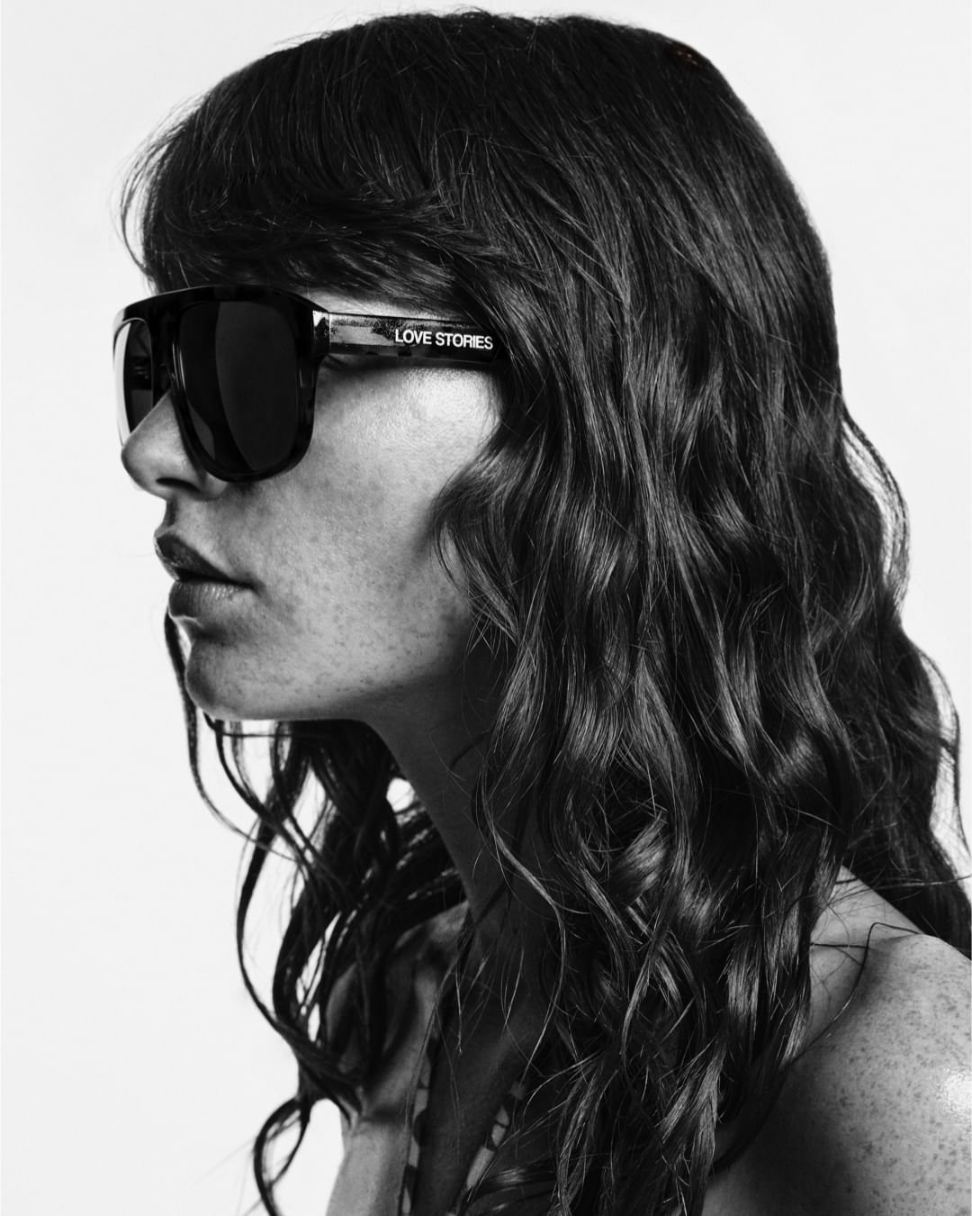Portrait of a woman wearing sunglasses with 'Love Stories' text on the side, shot in black and white with soft lighting.