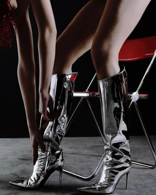 Woman's legs in shiny silver knee-high stiletto heel boots with a black background and a red metal chair create a fashion-forward studio shot.