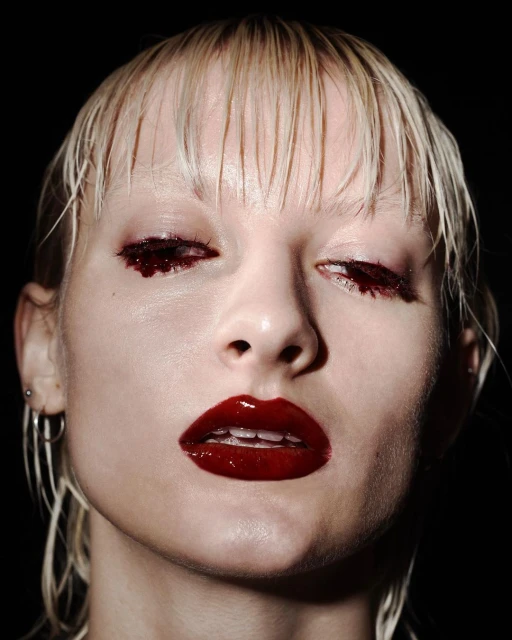 Close-up portrait of a pale-skinned adult woman with short blonde hair, dark red glossy lipstick, and dark red eyeshadow makeup.