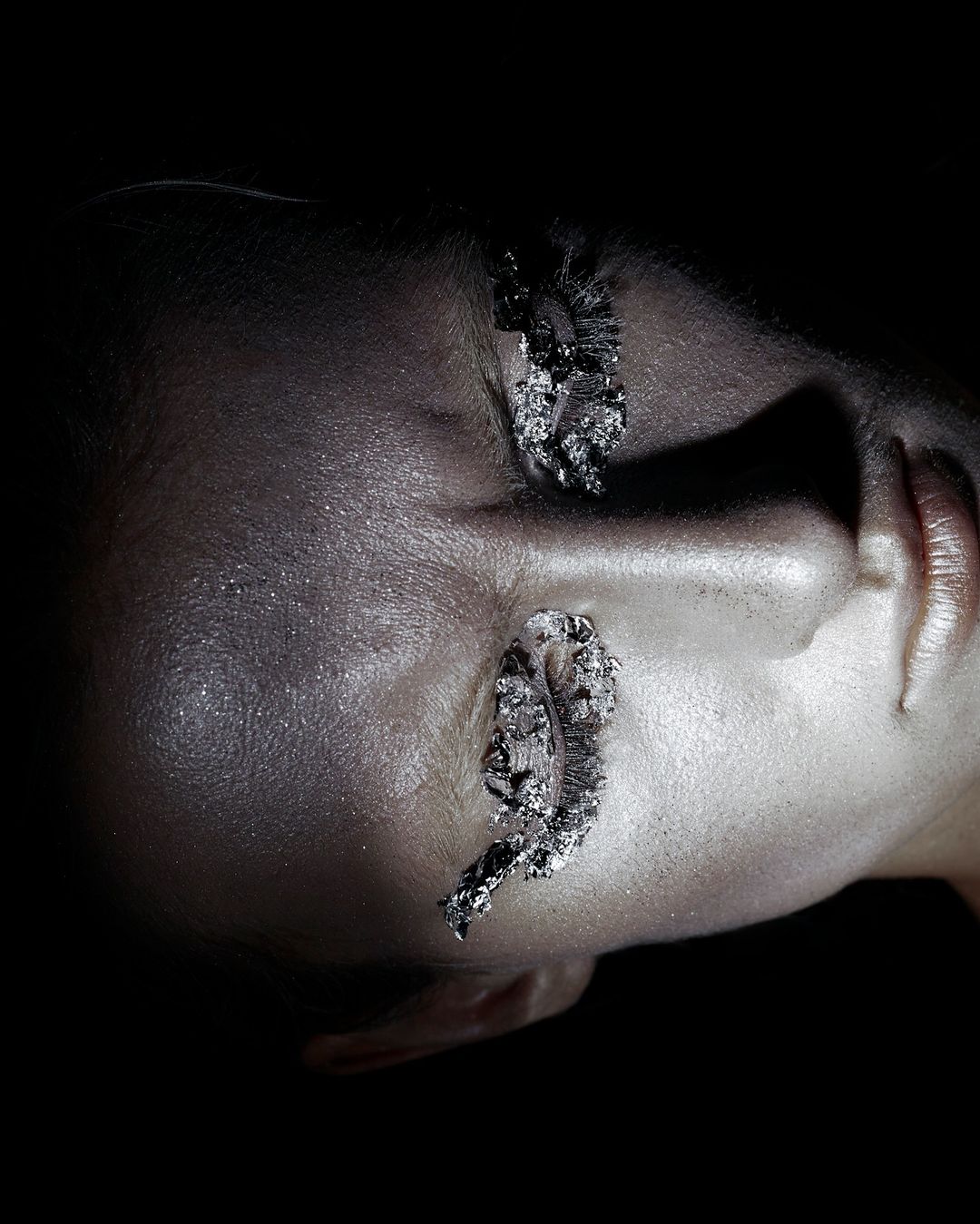 Close-up of a woman's face with silver metallic makeup, lips, and artistic eye decorations against a dark background in a beauty shot.