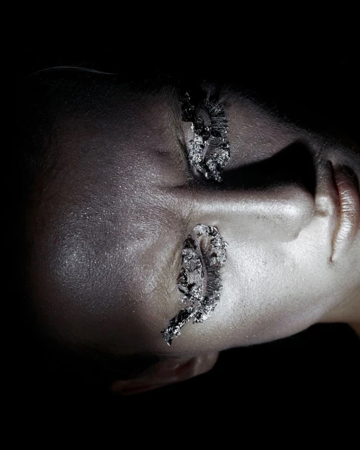 Close-up of a woman's face with silver metallic makeup, lips, and artistic eye decorations against a dark background in a beauty shot.