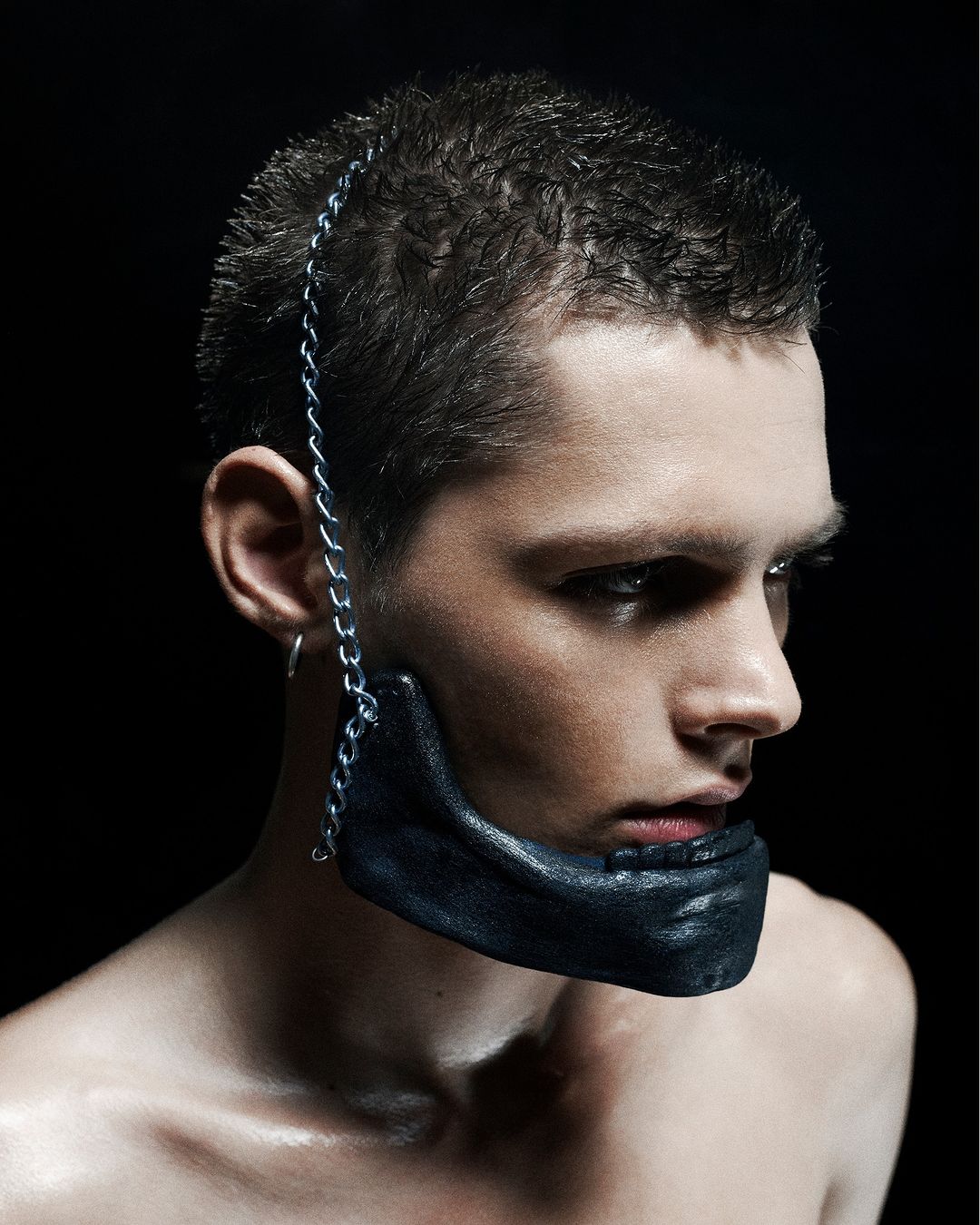 Striking studio shot of a young adult man with dark hair, bare shoulders, and a leather mandible mask secured by a silver chain.