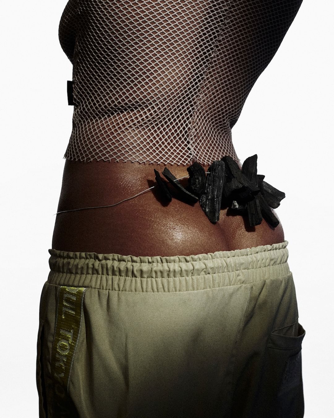 Close-up studio shot of person wearing beige pants, fishnet top, and black stone embellishment around waist, set against a white background.