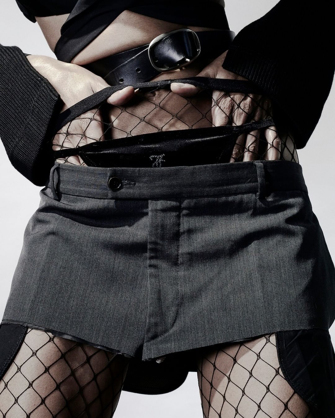 Torso shot of a fashion model in stylish grey shorts, a black top and fishnet stockings posing in a studio setting with a white background.