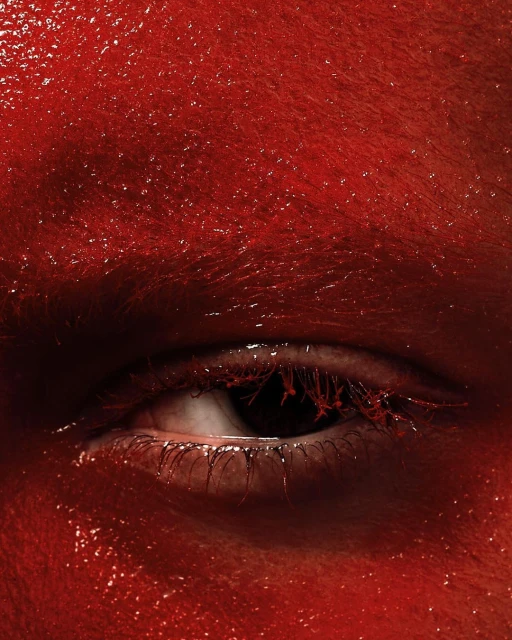 Macro shot of an eye and face covered in shiny red paint, creating a striking and textured visual.