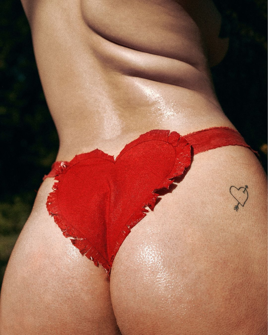Close-up of a woman's lower back and buttocks with a heart-shaped thong, featuring a tattoo of a heart pierced by an arrow.