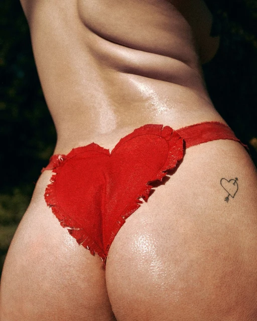 Woman with tanned skin poses outdoors wearing a red heart-shaped garment; a tattoo of a heart with an arrow decorates her skin.