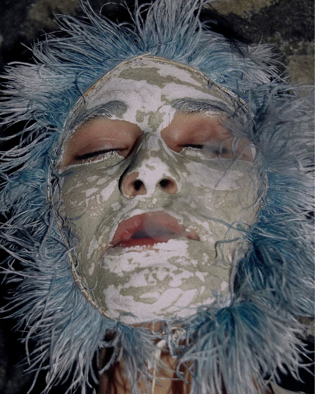 Close-up of a person's face with eyes closed, covered in a pale mask and surrounded by feathery blue headwear.