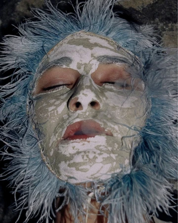 Close-up of a person's face with eyes closed, covered in a pale mask and surrounded by feathery blue headwear.