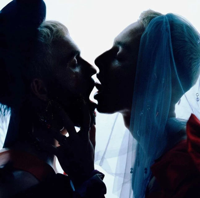 Two people in close proximity, possibly a couple, with blue veils or headwear in a dimly lit or shadowed environment, facing each other.