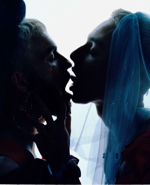 Close-up profile of two men kissing in dark tones, one with a sheer blue veil. Silhouetted figures with bright backlighting.