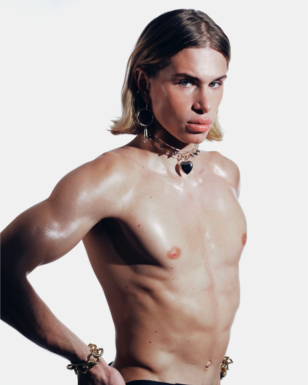 Glamorous shirtless adult male with blonde hair and jewelry stands in a bright white photo studio, showcasing fashion and beauty elements.