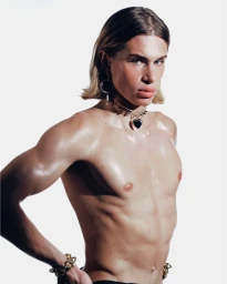 Shirtless person with blonde hair wearing a heart pendant choker and gold bracelets against a white seamless background