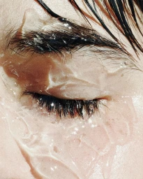 Close-up macro shot of a closed human eye covered in a glossy gel texture with scattered sparkles by Maud Ralliere