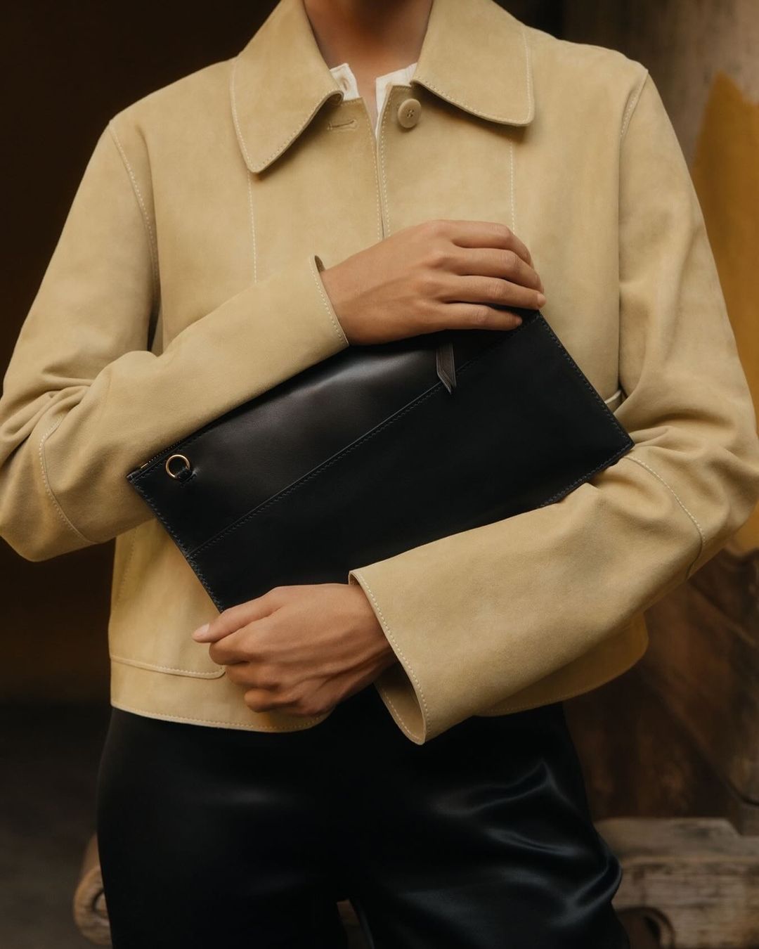 Model presents a black leather clutch bag wearing a beige suede jacket and black pants against a neutral background, fashion editorial.