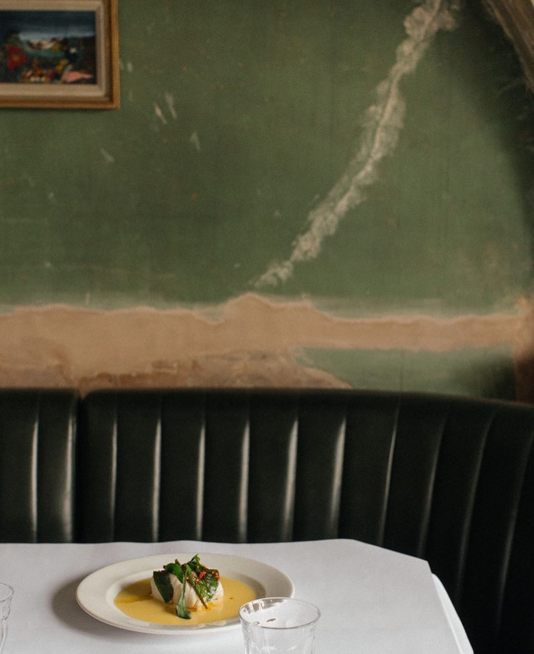Gourmet food plate on white linen-covered table in restaurant setting, with green wall and artwork in the background.