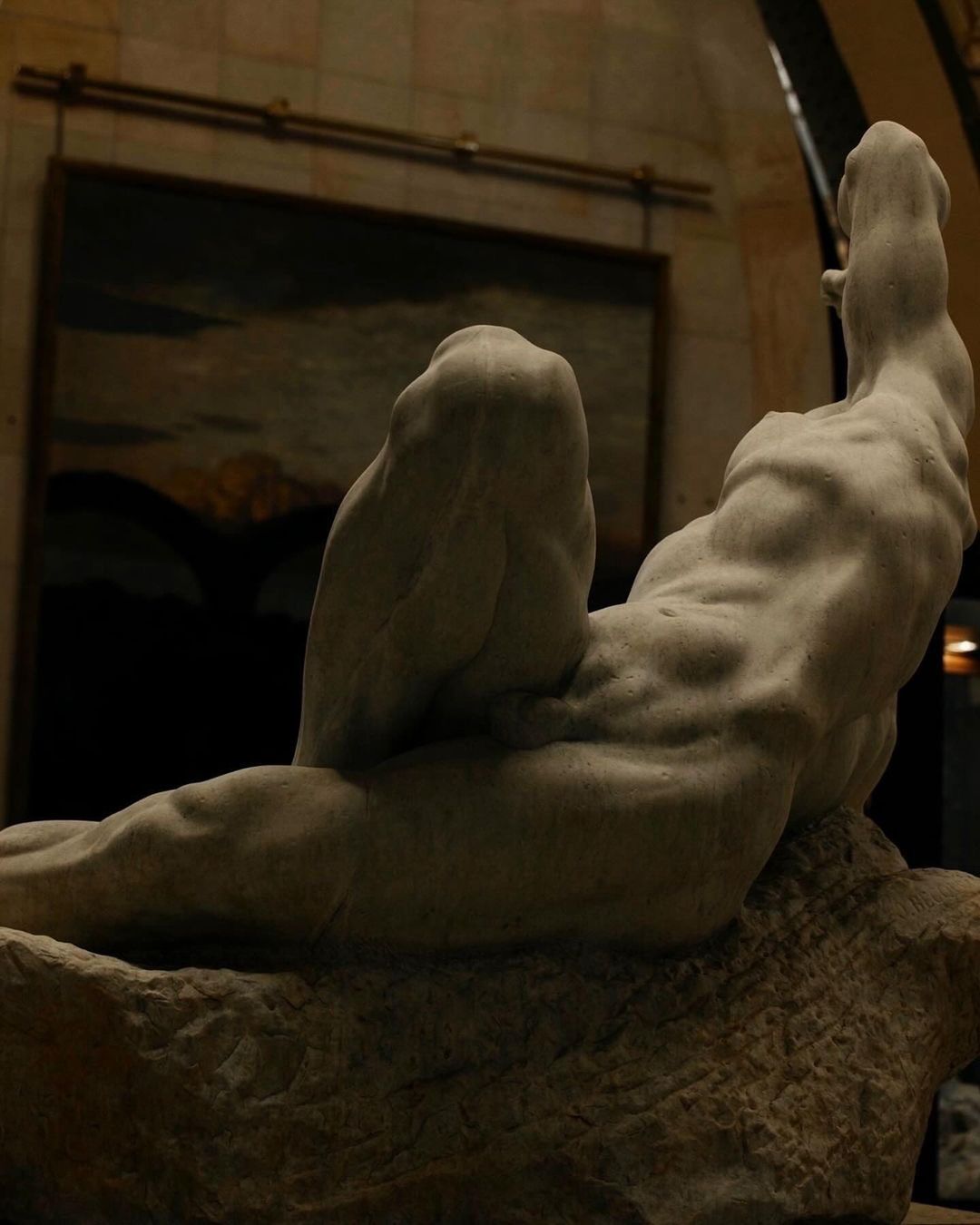 Stone sculpture of a reclining nude male figure indoors, backed by a dark painting and tiled wall, showcasing classical art and form.