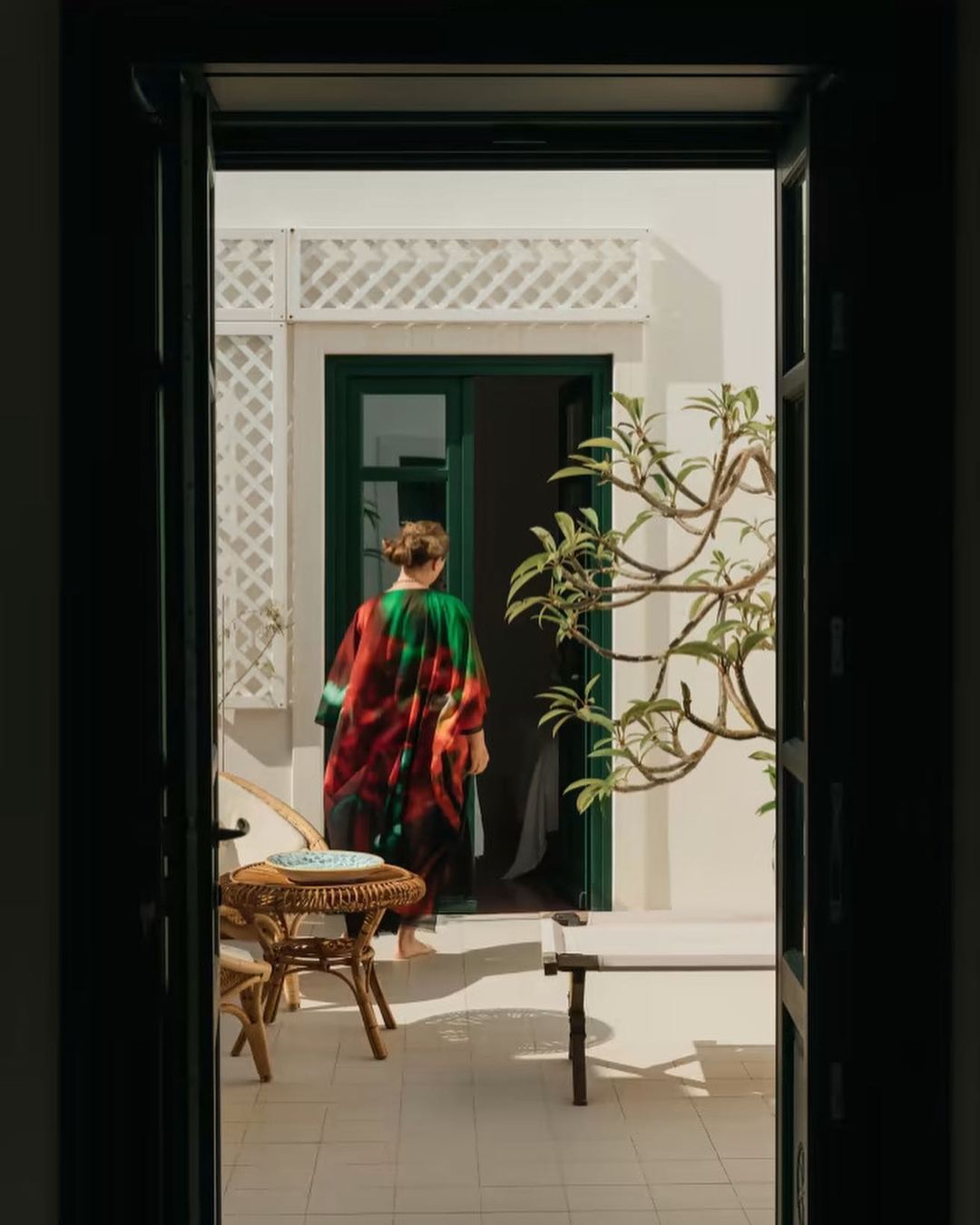 Woman walks through a doorway into a sunlit courtyard. Green door frame, white walls, potted plant, tile floor, table, chair, indoor/outdoor transition.