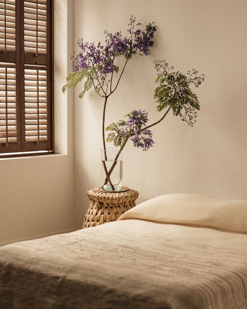 Bedroom interior with a cozy bed, woven side table, and vase holding purple flowers, light coming through shuttered window.