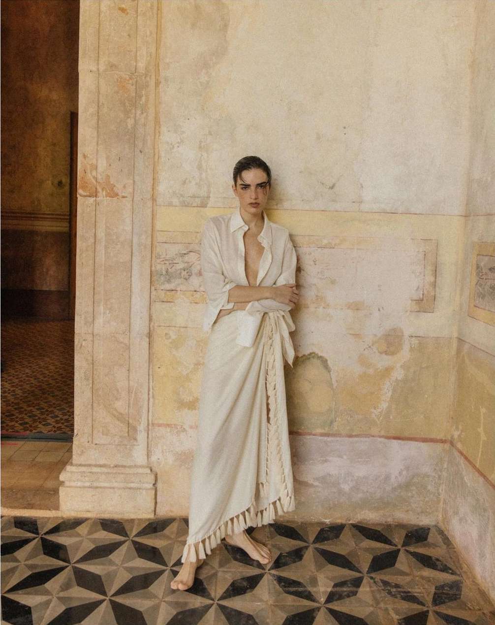 A woman in a white shirt and skirt poses against a textured wall with a geometric tile floor in an indoor setting, looking at the camera.