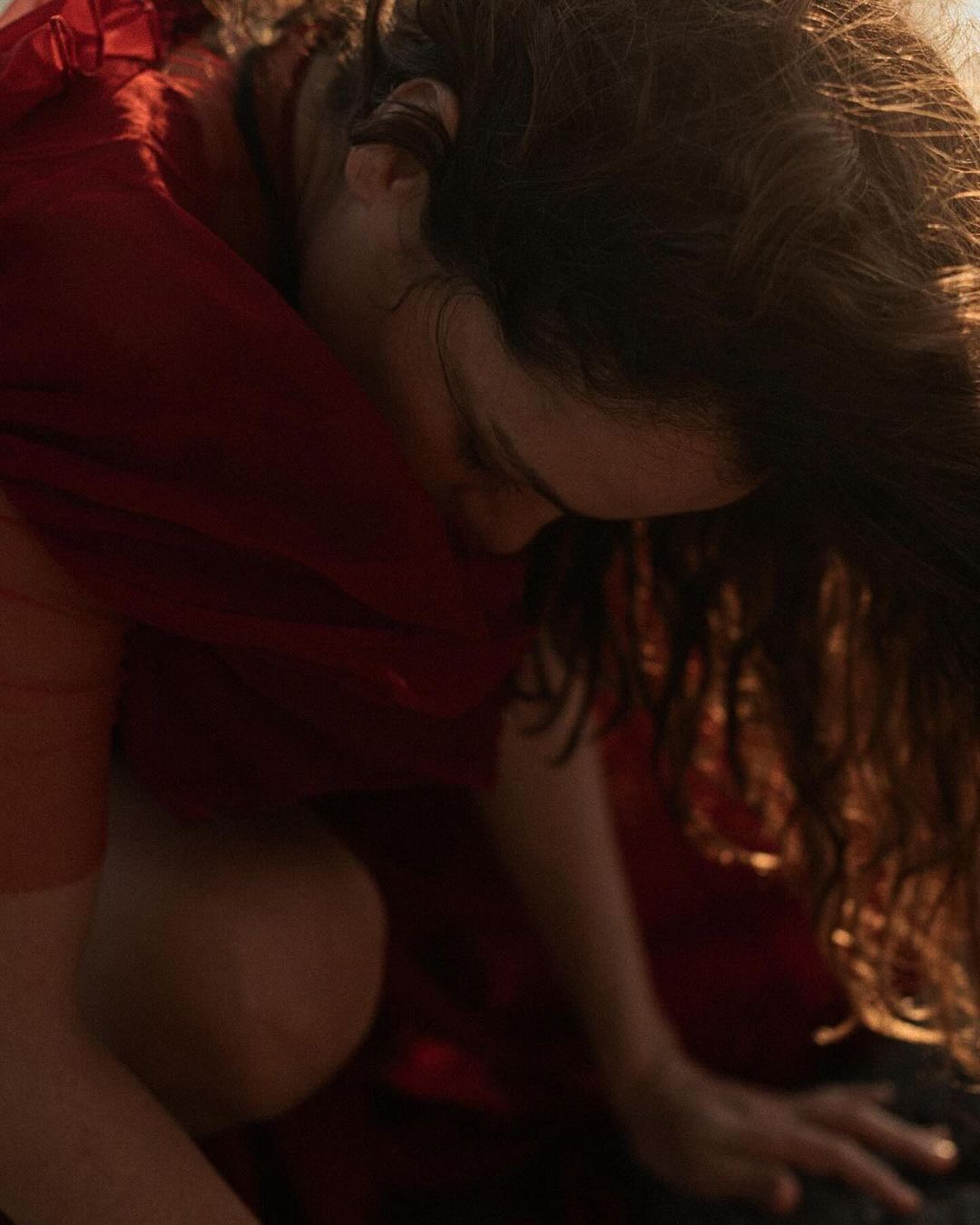 Indoor portrait captures a brunette woman in a red dress, crouching and looking down with soft, diffused lighting enhancing the scene.