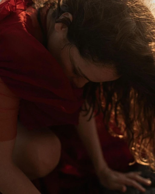 Indoor portrait captures a brunette woman in a red dress, crouching and looking down with soft, diffused lighting enhancing the scene.