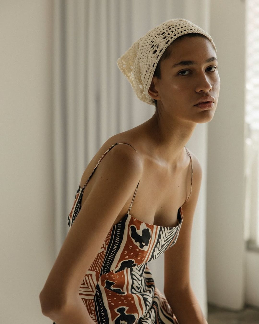 Elegant woman in a patterned sundress and crochet headscarf, posing indoors with soft, diffused light creating a fashion-editorial aesthetic.