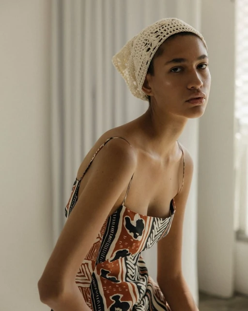 Elegant woman in a patterned sundress and crochet headscarf, posing indoors with soft, diffused light creating a fashion-editorial aesthetic.