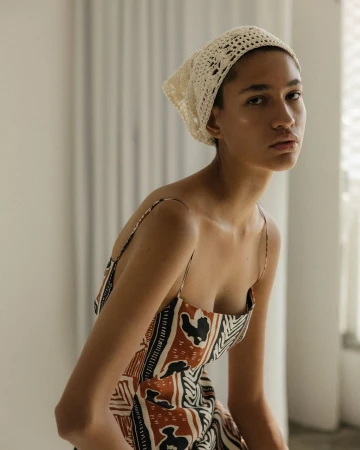Elegant woman in a patterned sundress and crochet headscarf, posing indoors with soft, diffused light creating a fashion-editorial aesthetic.
