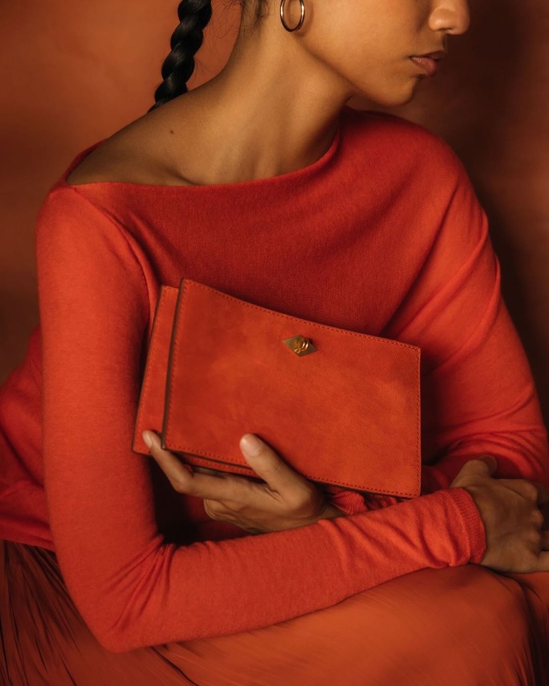 Adult female in a long-sleeve red dress holding a red clutch. The subject is set against a matching monochromatic orange background.