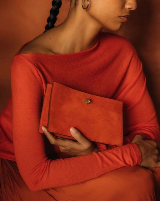 Adult female in a long-sleeve red dress holding a red clutch. The subject is set against a matching monochromatic orange background.