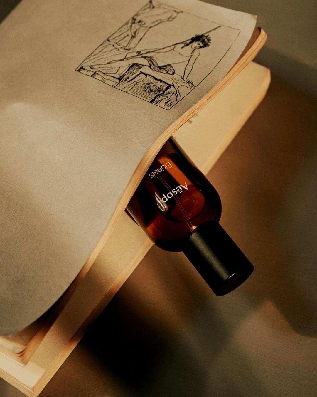 Aesop bottle nestled in an open book featuring a black and white drawing, close-up product shot with neutral tones and textured surfaces.