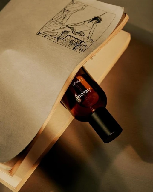 Aesop bottle nestled in an open book featuring a black and white drawing, close-up product shot with neutral tones and textured surfaces.