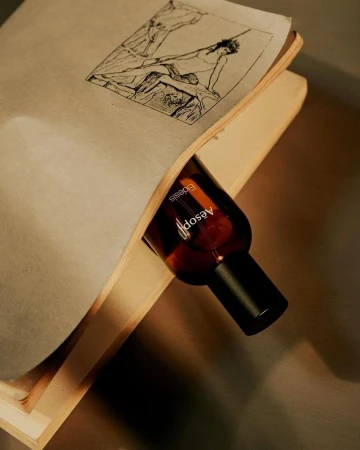 Aesop bottle nestled in an open book featuring a black and white drawing, close-up product shot with neutral tones and textured surfaces.