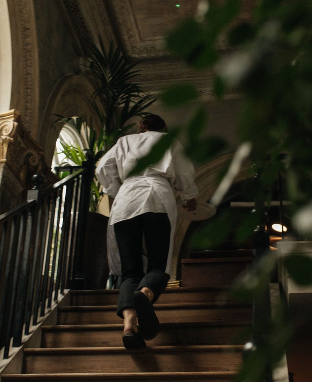 Back view of a woman in a white jacket and black pants walking up wooden stairs inside a building with a railing and potted plants.