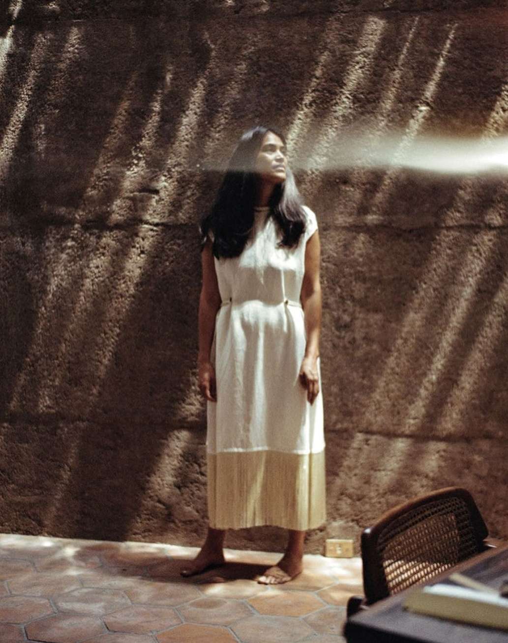 A woman in a white dress stands against a textured wall with dappled sunlight, with a chair and tiled floor adding to the scene's aesthetic.